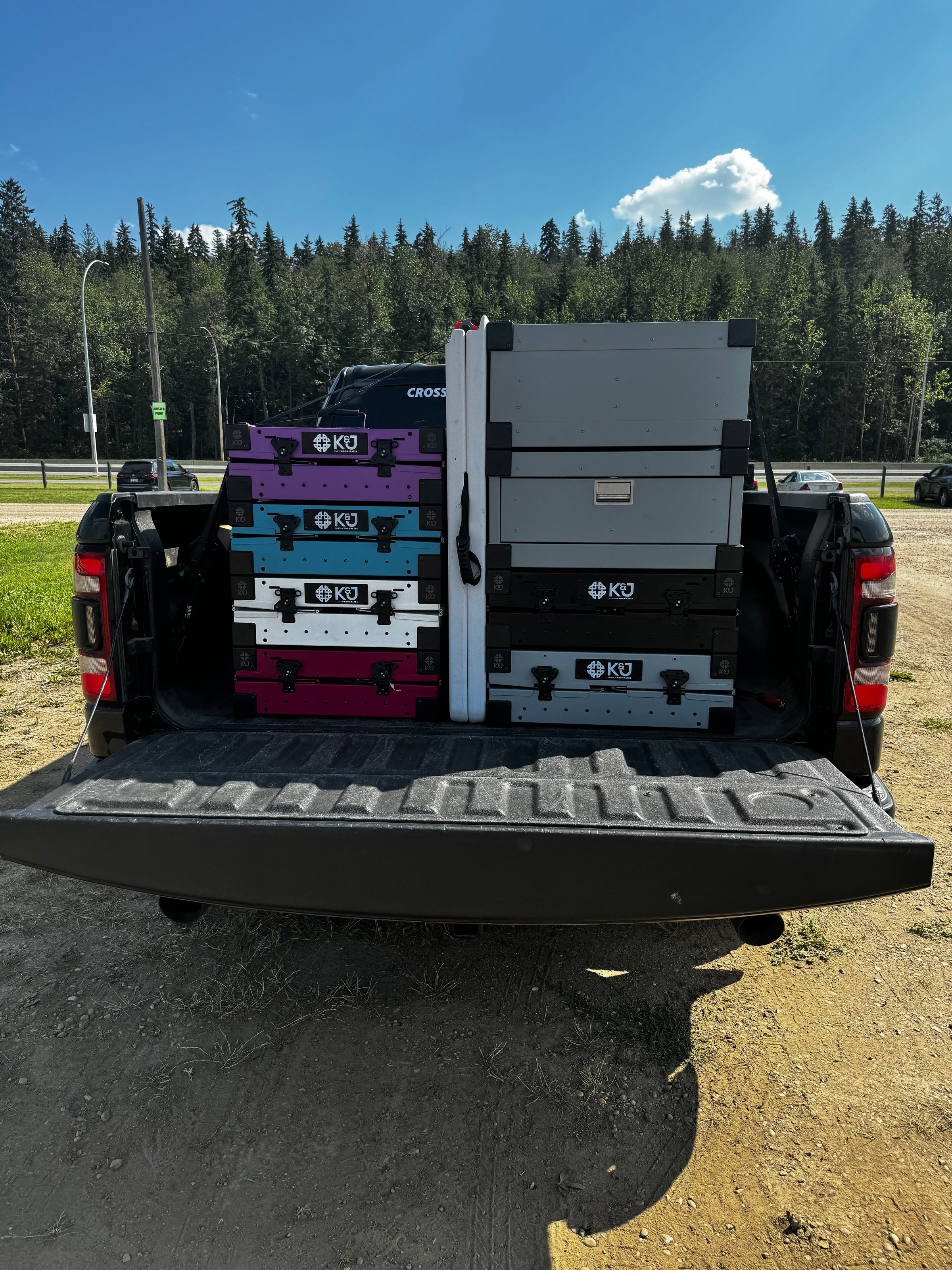 Black Dodge Ram truck with tailgate open, showing 7 collapsed K&J dog crates stacked for easy storage. Crates in purple, teal, white, pink, grey, and black colors. Truck parked in a green field for efficient transport.