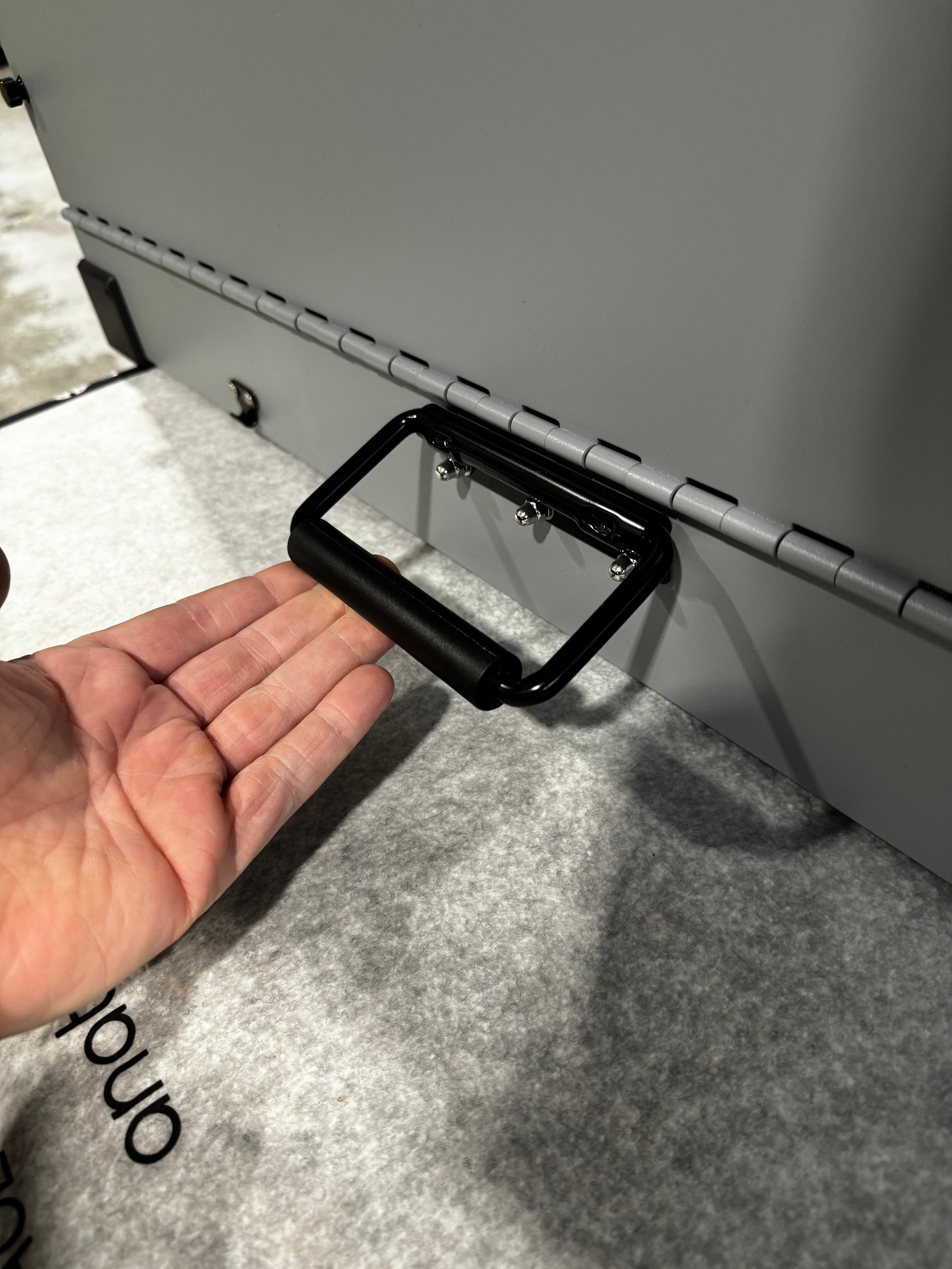 Large grey collapsible K&J crate in an industrial warehouse, with a close-up of the spring-loaded side handle being held out by a person's hand.